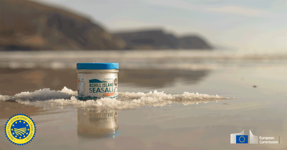 Image showing a tub of Achill Island sea salt against the background of an Achill Island beach and featuring the Protected Geographical indication and EU logos