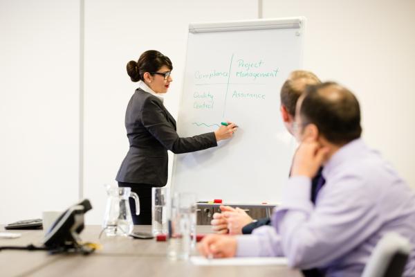 Woman standing at whiteboard with two men seated