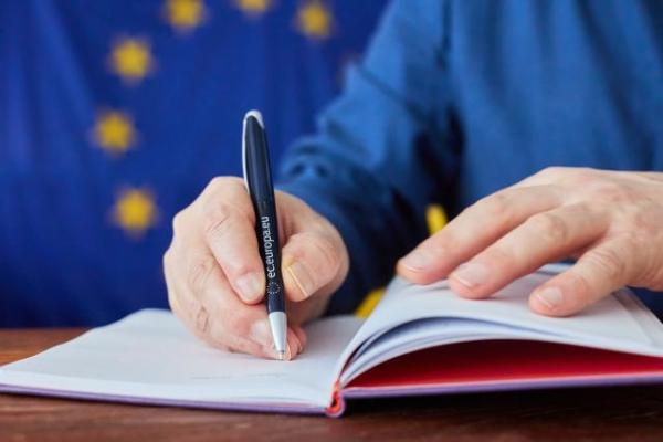 Person writing in a notebook with an EU pen and the EU flag in the background
